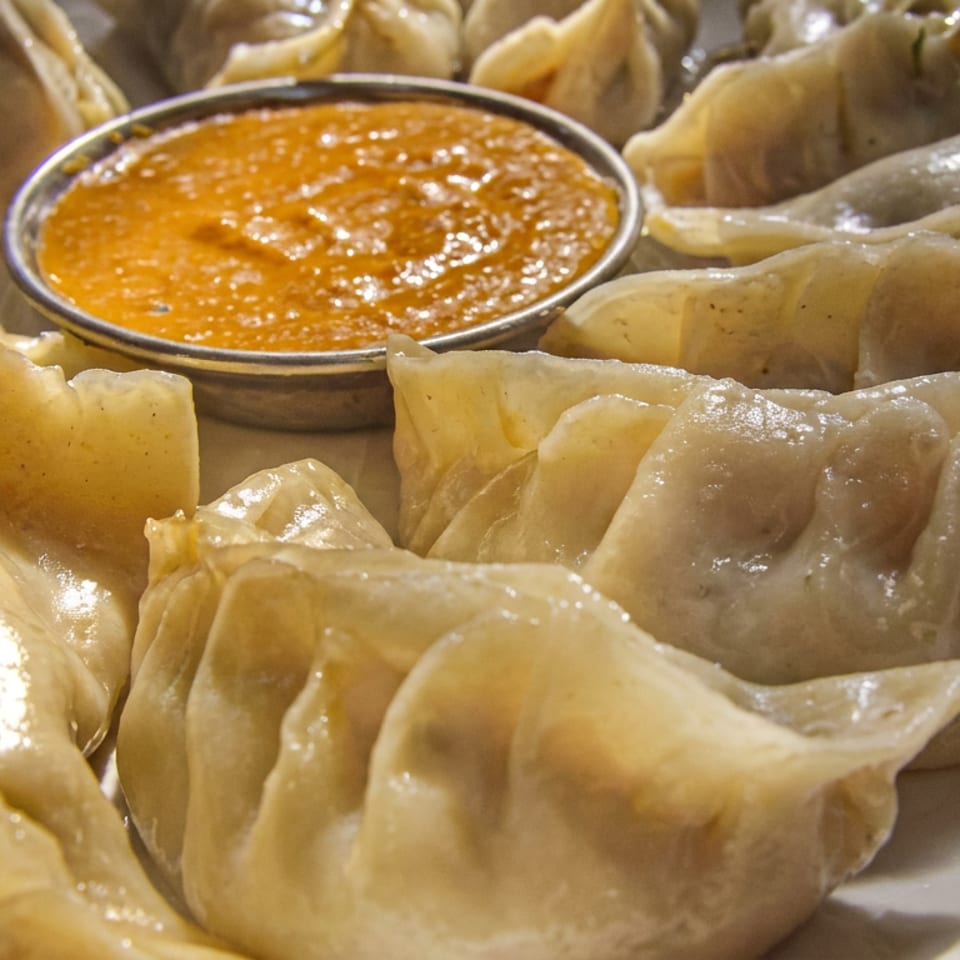 A plate of steamed momos, a type of dumpling, surrounds a small bowl of orange dipping sauce.