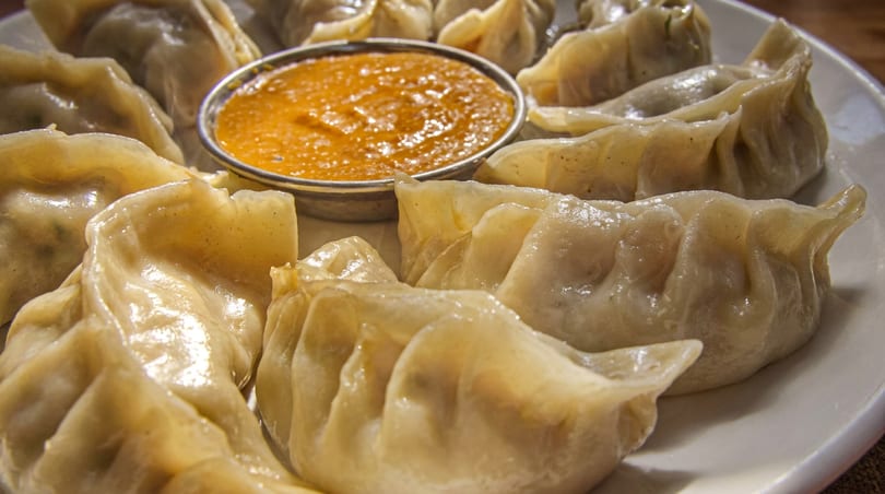 A plate of steamed momos, a type of dumpling, with a small bowl of orange dipping sauce.