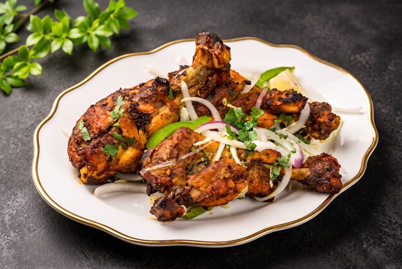 Spicy tandoori chicken drumsticks and thighs, garnished with onions, cilantro, and lemon on a white plate.