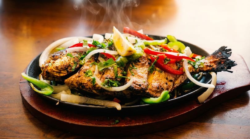 Steaming grilled whole fish with lemon, onions, and peppers on a sizzling cast iron platter.