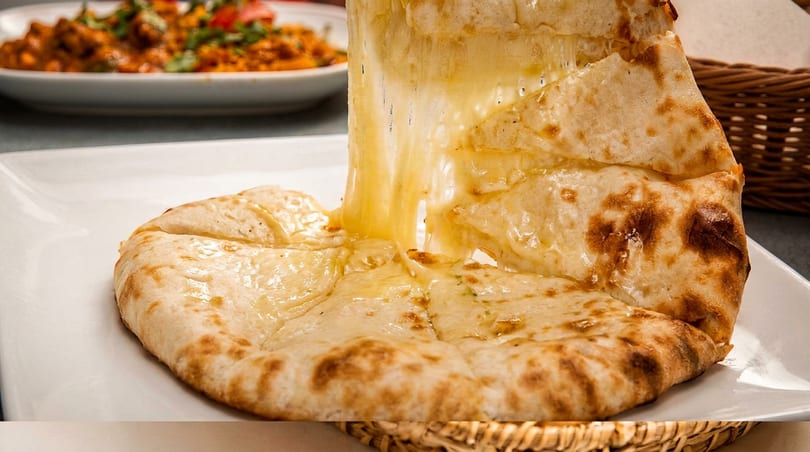 Cheesy naan bread being pulled apart, showing melted cheese stretching, with a curry dish in the background.