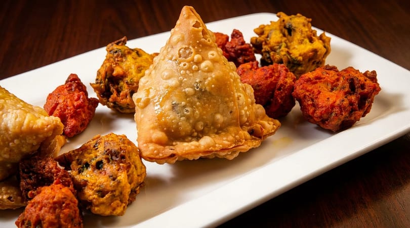 A white plate holds golden samosas and vibrant red and yellow pakoras, a delicious appetizer assortment.