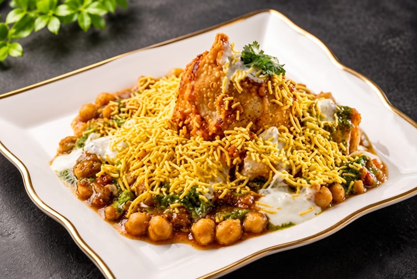 Samosa chaat with chickpeas, sauces, and crispy sev on a white plate with a gold rim.