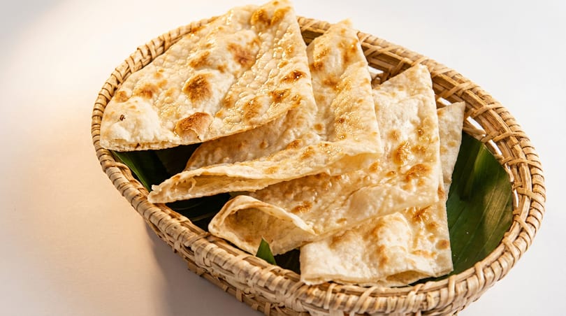 Golden brown flatbread pieces, possibly naan or roti, served in a woven basket lined with a green leaf.