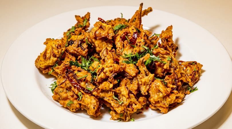 A white plate piled high with golden-brown pakoras, garnished with fresh green herbs and dried red chilies.