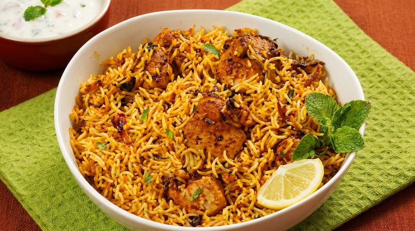 A white bowl of chicken biryani with mint and a lemon wedge, alongside a small bowl of raita.