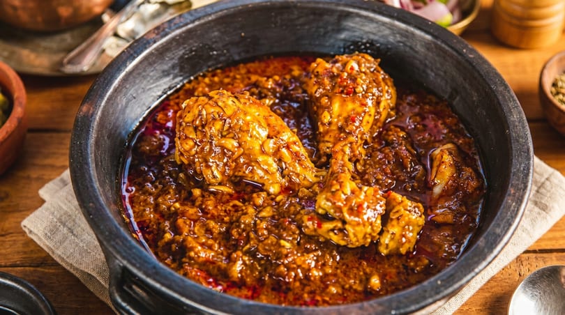 Chicken curry with drumsticks in a black bowl, surrounded by spices and a spoon on a wooden table.